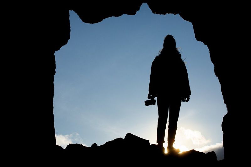 Sagoma di una persona con una macchina fotografica, in piedi davanti all'ingresso di una caverna, che si staglia contro un cielo soleggiato e luminoso.