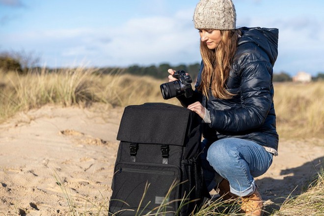Una donna accovacciata su una spiaggia sabbiosa, mentre ritira una fotocamera Canon nella borsa del kit.