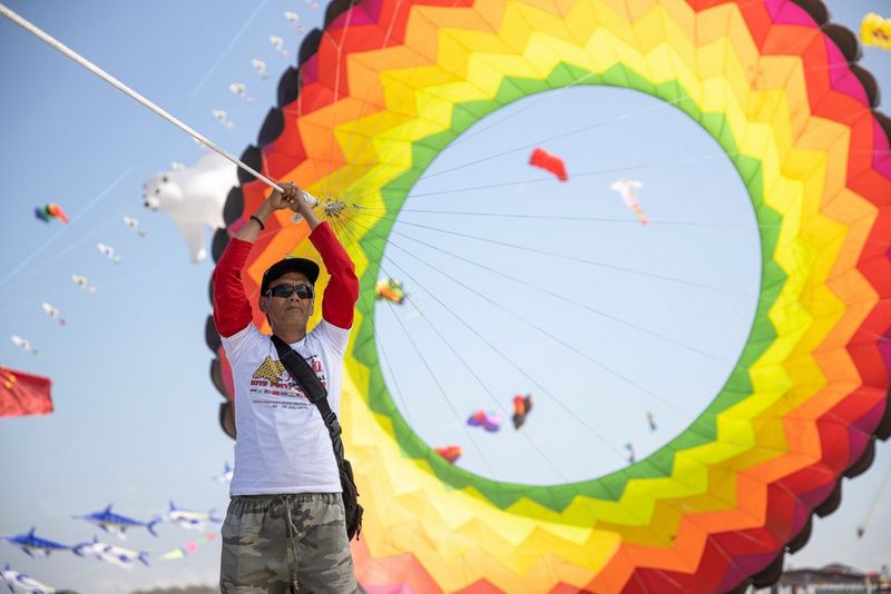 A portrait of a person holding the strings of a large, rainbow-coloured ring kite, with blue sky and other kites visible behind it, taken with a Canon RF 24-70mm F2.8L IS USM lens.