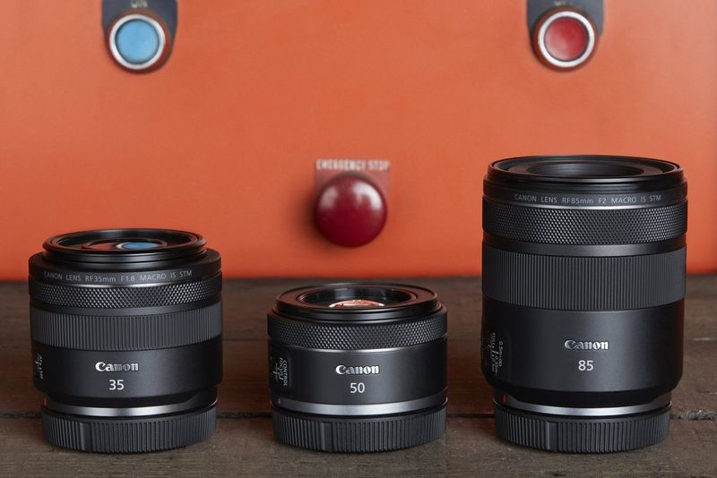 Three Canon RF lenses – RF 35mm F1.8 MACRO IS STM, RF 50mm F1.8 STM and RF 85mm F2 MACRO IS STM – on a wooden surface with an orange wall behind.