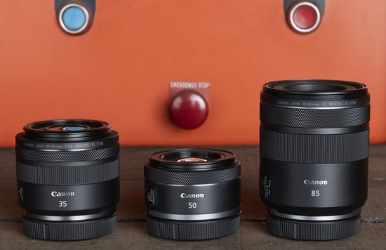 Three Canon RF lenses – RF 35mm F1.8 MACRO IS STM, RF 50mm F1.8 STM and RF 85mm F2 MACRO IS STM – on a wooden surface with an orange wall behind.
