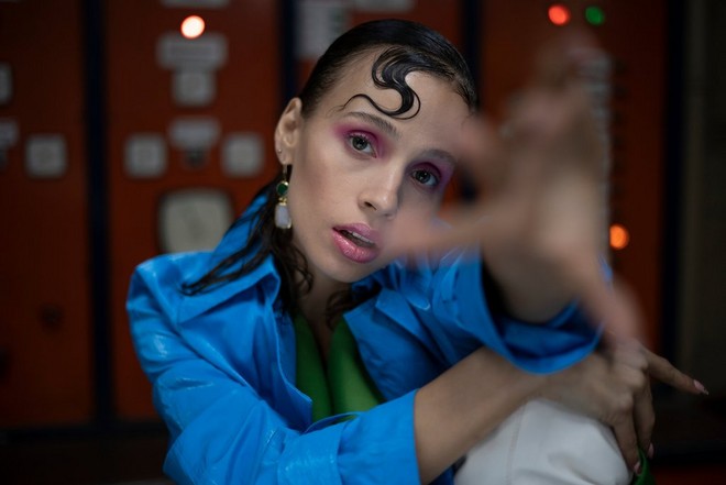 A shot of a woman wearing a blue coat and bright pink eyeshadow, reaching her hand towards the camera.