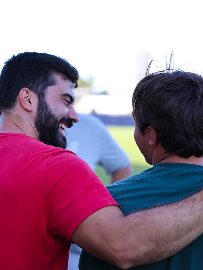 One man puts his arm around another, while they smile at one another on a training pitch