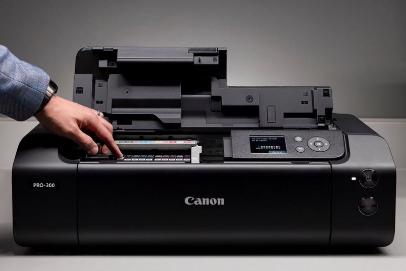 Верхняя крышка принтера Canon imagePROGRAF PRO-300 открыта, а человек рукой устанавливает один из 10 чернильных картриджей.