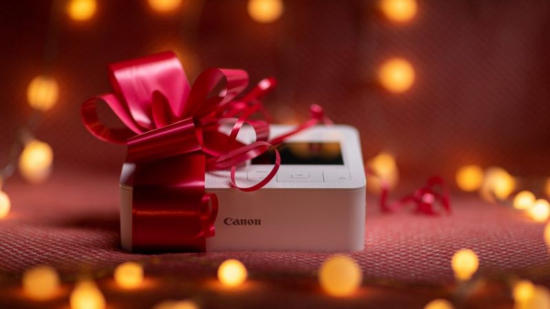 Canon SELPHY CP1500, mis on pakitud punase paelaga ja ümbritsetud häguste jõulutuledega.