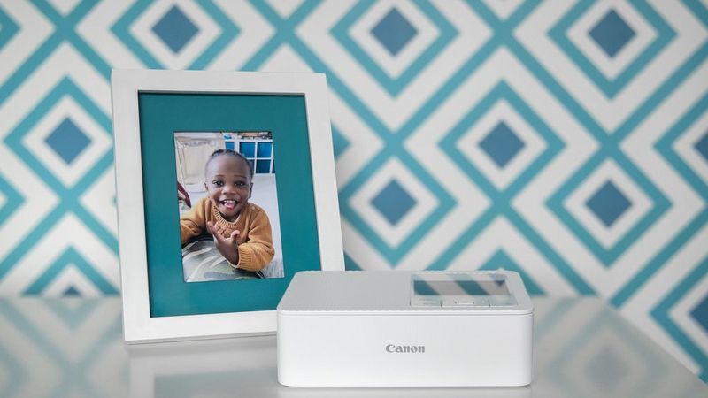 White SELPHY CP1500 printer on a white surface with a picture of a little girl in a frame standing behind it.