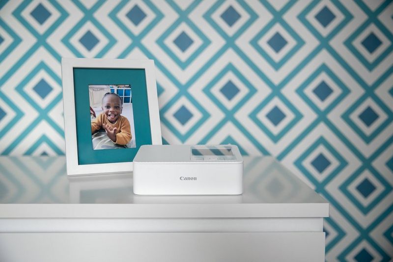 Бел печатач SELPHY CP1500 врз бела комода со фиоки, врз неа слика од мало девојче кое се смее, ставена во бела рамка покрај печатачот. Во заднина има ѕидни тапети со сина графика.