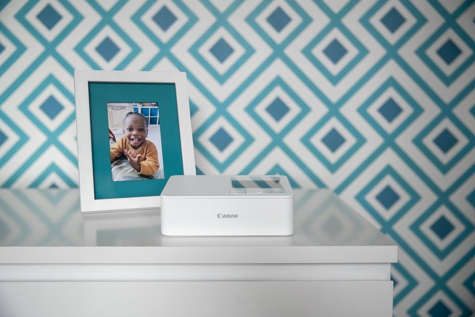 Una SELPHY CP1500 en blanco sobre una cómoda blanca con una imagen de una niña sonriendo en un marco blanco junto a ella. Un fondo de pantalla azul gráfico al fondo.