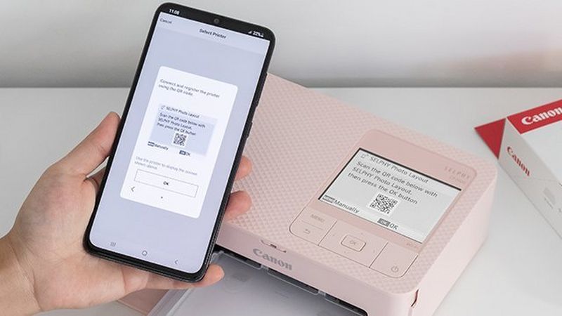 A hand holding a phone scanning a QR code from the pink SELPHY CP1500 printer LCD screen. SELPHY paper packaging is visible in the right corner.