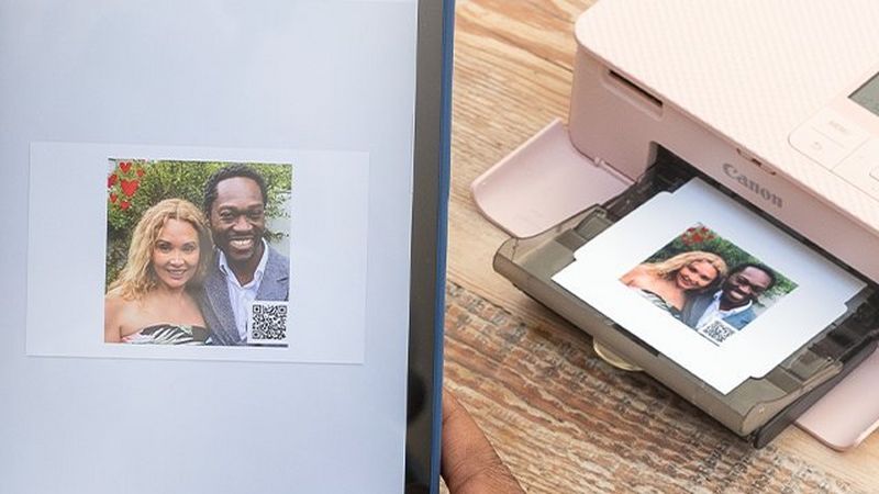 A tablet with a picture of a couple in SELPHY Photo Layout app with a QR code in the bottom right of the picture. SELPHY CP1500 in pink in the background with the same picture printed out.