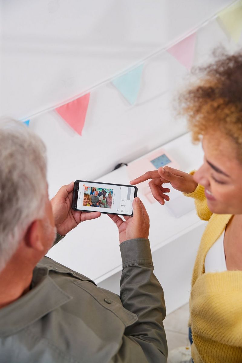 Deux mains tenant un téléphone avec une photo de famille, sur laquelle des tampons ont été appliqués à l'aide de l'application SELPHY Photo Layout. Une autre main dirigée vers le téléphone est visible dans le coin supérieur droit.