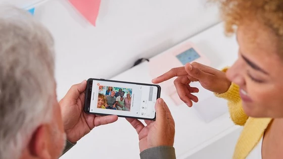 Две раце држат телефон со слика од семејство, а врз сликата има печати што се додаваат со апликацијата SELPHY Photo Layout. Во горниот десен агол се гледа друга рака која покажува кон телефонот.