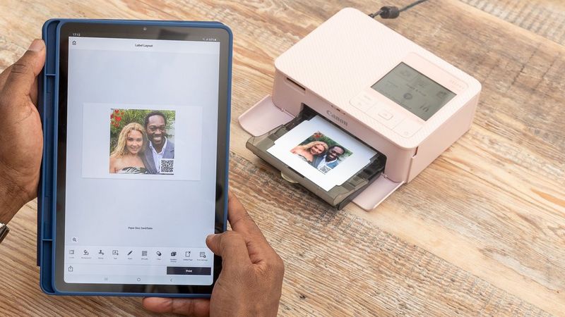 Une tablette dont l'écran affiche la photo d'un couple dans l'application SELPHY Photo Layout avec un code QR en bas à droite de l'image. En arrière-plan, une imprimante SELPHY CP1500 rose imprime la même image.