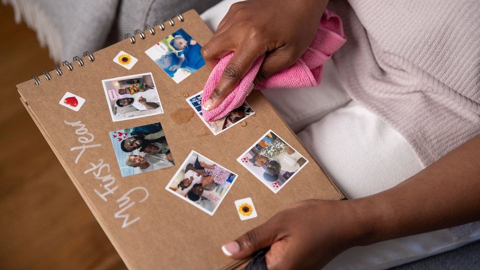Una mujer tiene en su regazo un álbum de fotos de recortes titulado «Mi primer año» con imágenes de una niña pequeña sola o con sus padres y limpia gotas de agua de una de las fotografías con un paño rosa brillante.