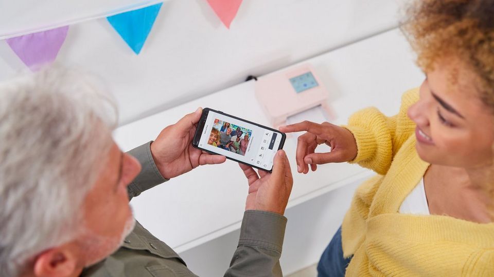 Un hombre mayor sostiene un teléfono con una imagen de una familia con sellos aplicados en la aplicación SELPHY Photo Layout. Una joven junto a él señala el teléfono. Se puede ver la SELPHY CP1500 en rosa en el fondo.