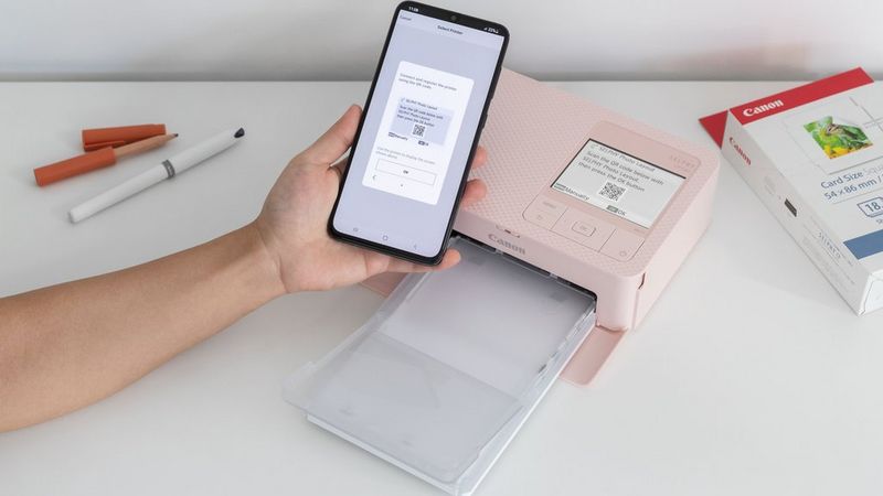 Рака која држи телефон што скенира QR-код од LCD-екранот на розовиот печатач SELPHY CP1500. Во десниот агол се гледа пакување хартија SELPHY и пенкало, а во левиот агол се гледа молив.