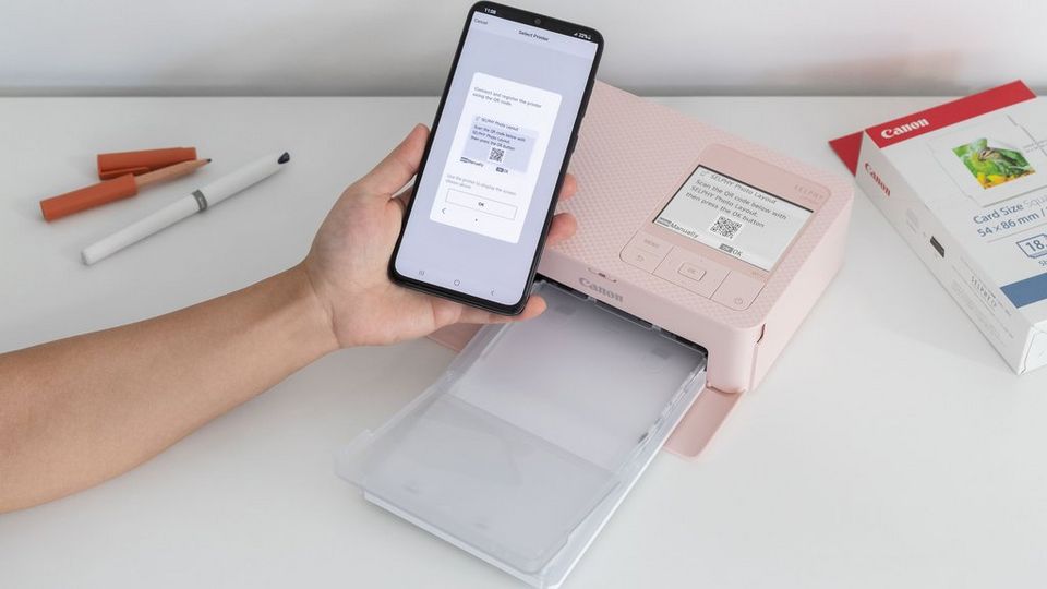 Una mano con un teléfono escanea un código QR desde la pantalla LCD de la impresora SELPHY CP1500 en rosa. Se pueden ver el embalaje de papel de la SELPHY en la esquina derecha, y un bolígrafo y un lápiz en la esquina izquierda.
