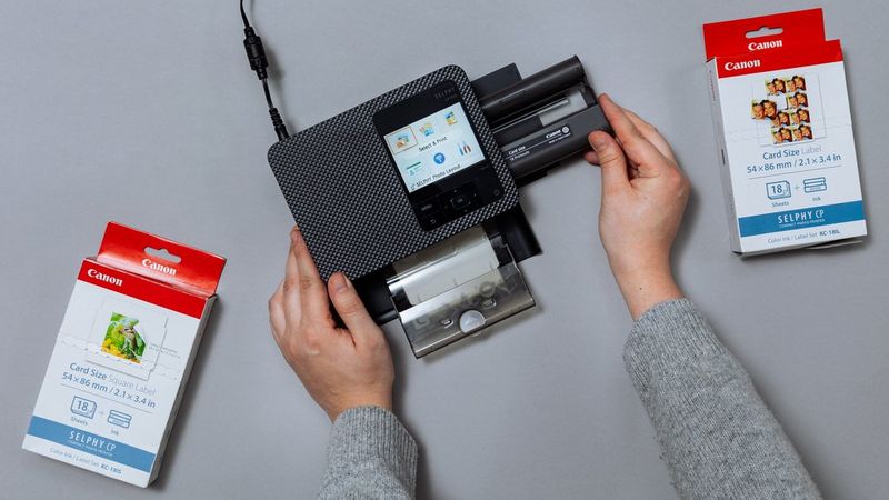 A person putting in the ink into the black SELPHY CP1500 on a table with packages of print paper around the printer. 