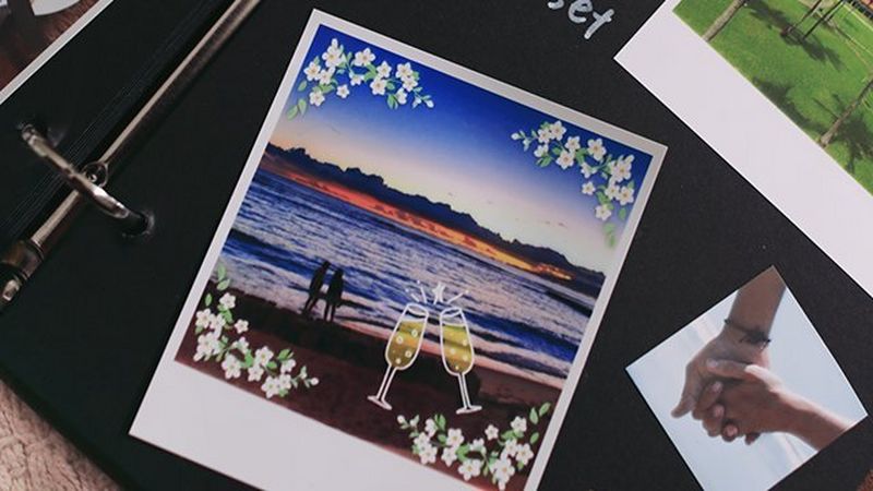 A square print stuck in a scrapbook with a picture of sunset at the beach with a silhouette of two people and a flower frame applied. Next to it a small square image of two holding hands.