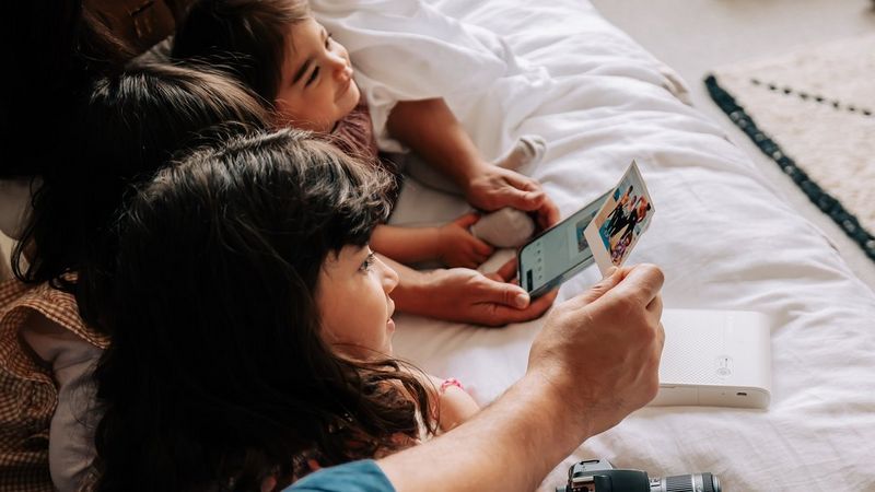 Three young children sat on a bed together looking at printed family photos.