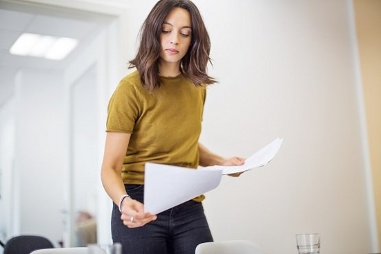 Woman with brown hair and wearing a yellow top carries paper in both hands 