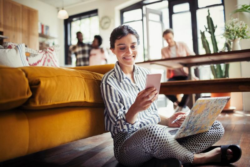 An image of a smiling woman using a laptop and texting with a smartphone