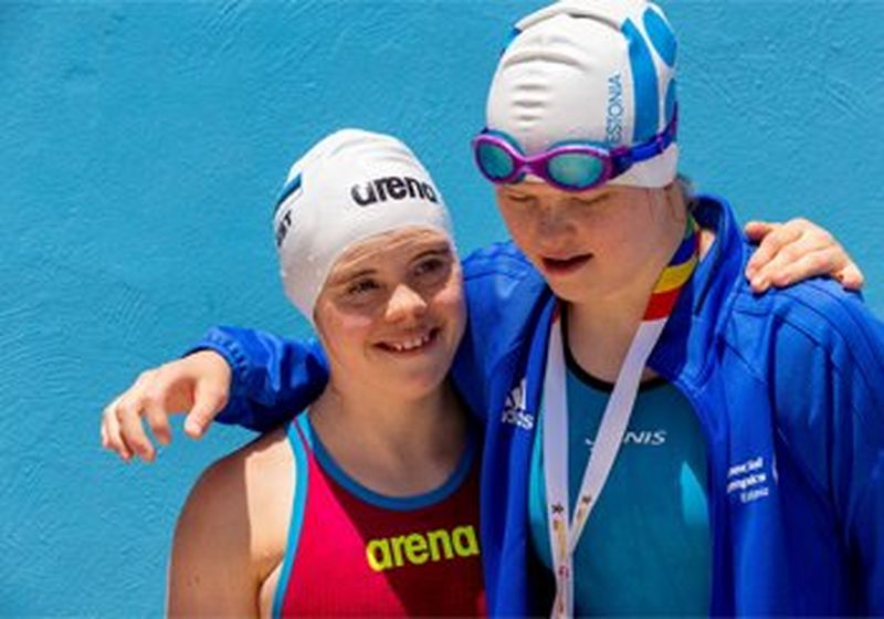 Two swimmers photographed from the chest up. The swimmer on the right has their arm around the shoulders of the swimmer on the left. They are both wearing white swimming caps, with goggles pushed up over it on the right-hand swimmer. She also wears a blue jacket over a blue swimming costume and the ribbon of a medal can be seen around her neck. The left-hand swimmer wears a red costume with blue trim.