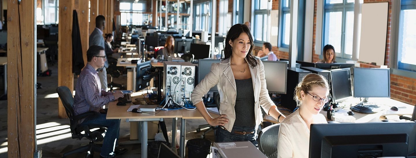 Woman with hand on her hip stands over another woman working at a large computer screen in an office warehouse setting.