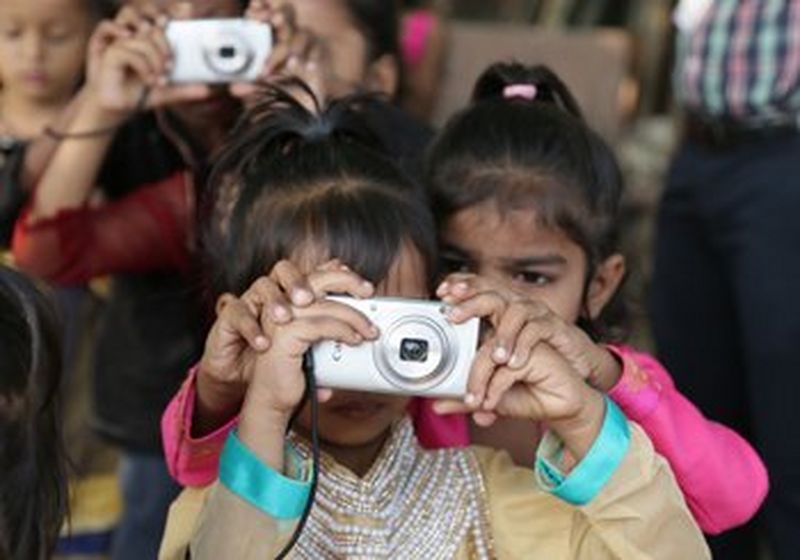 A little girl holds a Canon camera up to her face to take a picture, while behind her another little girl places her hands over hers and looks over her shoulder.