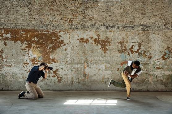 Photographer David Newton crouches down to capture a dancer mid-move against a distressed concrete wall.
