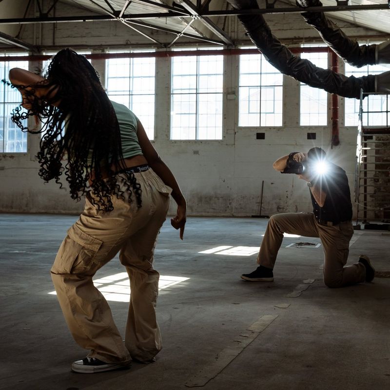 A photographer crouches down to photograph a woman dancing in a warehouse, his face obscured by the flare created as the Canon Speedlite EL-5 fires.