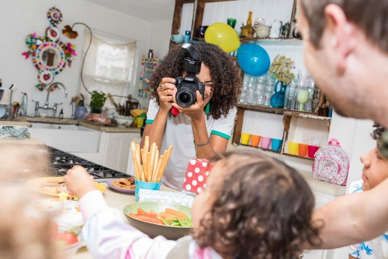 A young woman is taking a photo with a camera and a Speedlite flash pointing sideways and upwards of a toddler sitting by the table surrounded by their family.