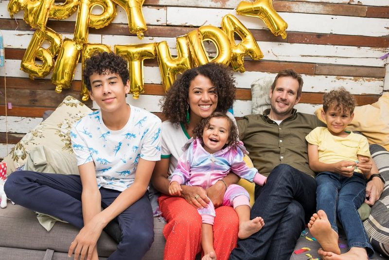 A portrait of a family with parents and 3 children, all sitting on a couch with balloons spelling happy birthday on the wall behind them. 