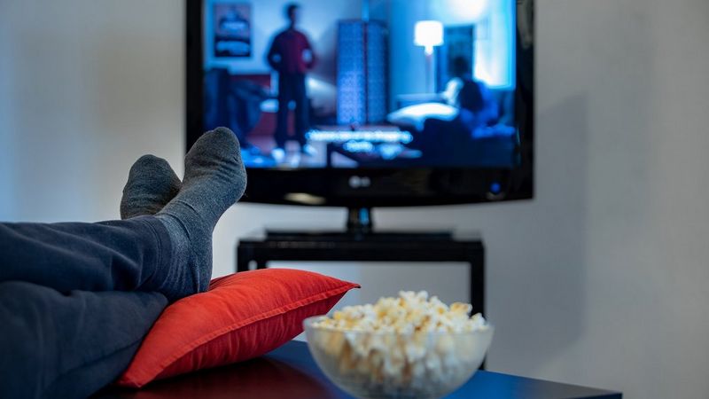A pair of socked feet rest on a red cushion on a table. Beside them is a bowl of popcorn. Beyond both is a blurred TV playing a show with subtitles.