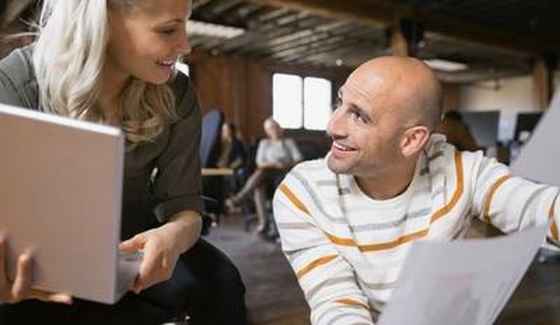 Blonde-haired woman rests a laptop in her hands as she chats to a bald man dressed in a stripy white jumper.