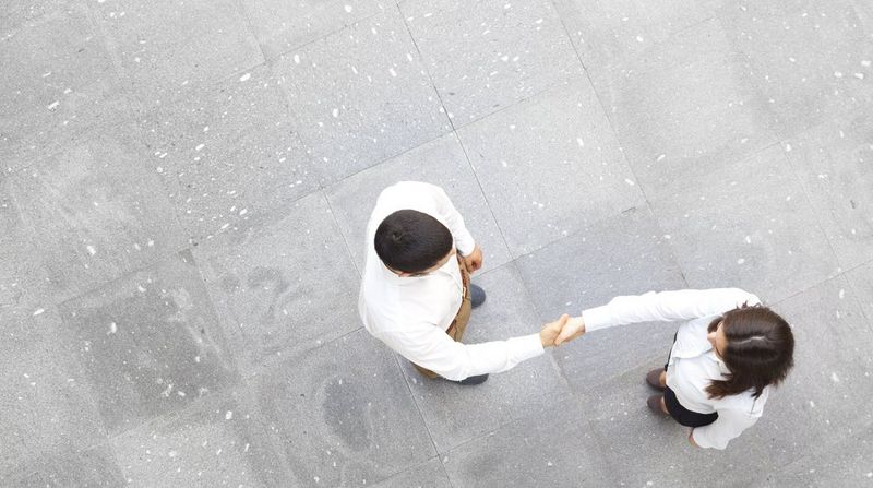 Two white-shirted business professionals photographed from above as they shake hands on a grey tiled floor.