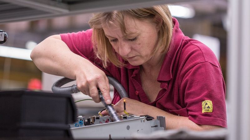 A blonde woman in a burgundy polo shirt is photographed through industrial racking. She is using a tool to clean a piece of electronic equipment, housed in a metal box.