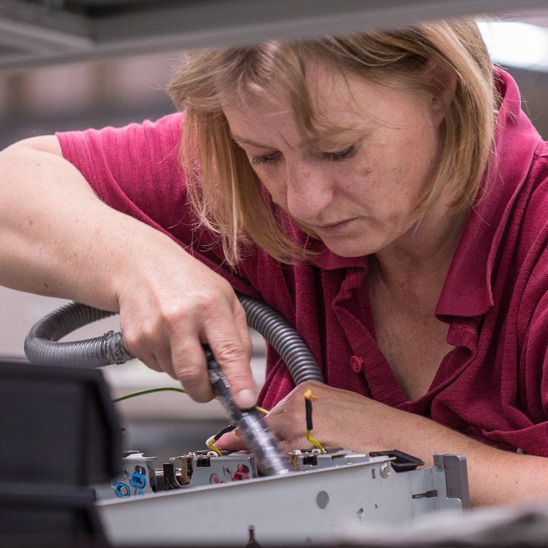 A blonde woman in a burgundy polo shirt is photographed through industrial racking. She is using a tool to clean a piece of electronic equipment, housed in a metal box.