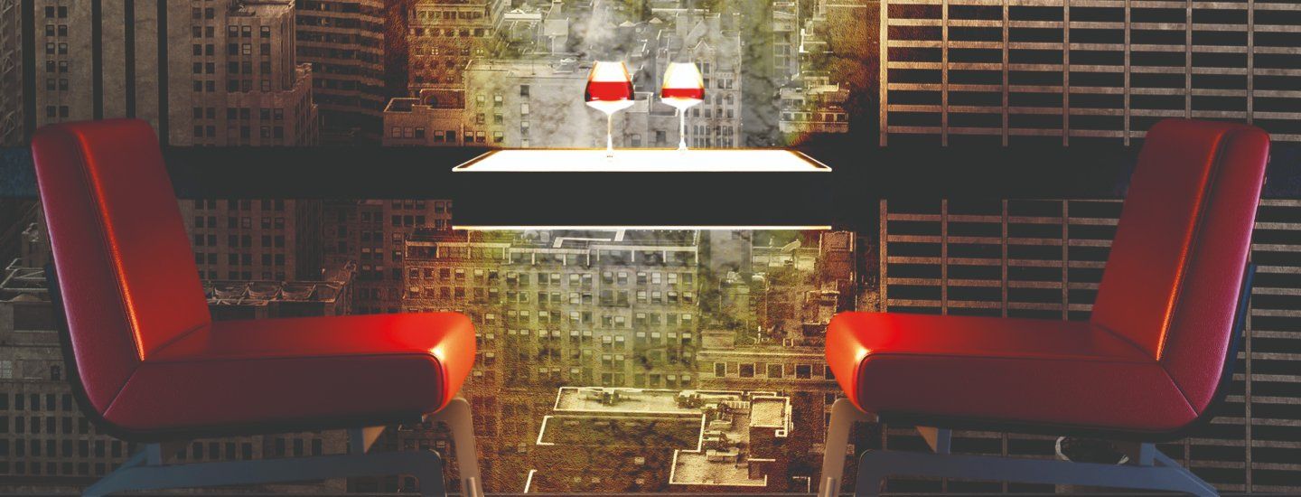A table set with two wine glasses flanked by two red chairs in front of digitally printed wallpaper showing a cityscape