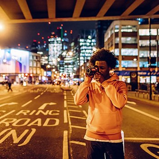 A man stands under a city centre bridge beside a road to take a photo with a Canon camera.