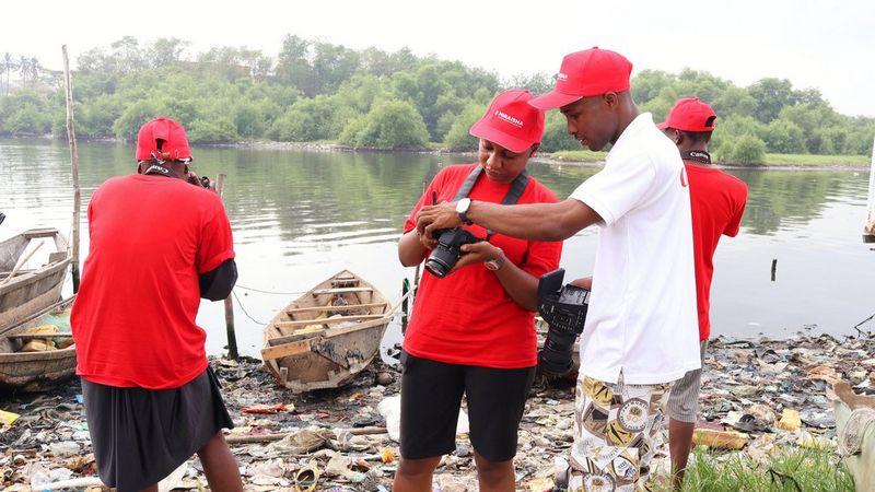 Four people stand beside a lake and the ground is covered in detritus. To the fore of the photo, a young man in a white shirt stands with a person of undefined gender and they both look into the back screen of the camera they hold.