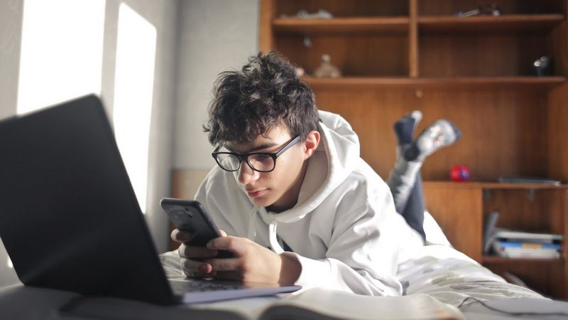 A teenaged boy wearing glasses and a white hoodie lies on his stomach on his bed with his laptop open in front of him. He holds his phone in both hands and checks it.