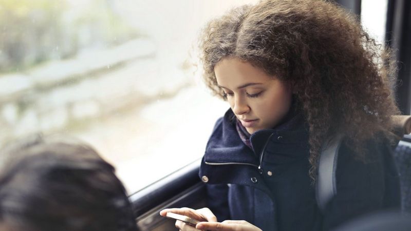 A teenaged girl with long, dark afro hair and wearing a winter coat sits on a bus and checks her phone.
