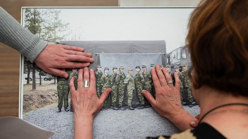 Photographed from behind. the head and shoulders of a woman as she lays her hands on a tactile print of a group of people wearing army camouflage. A third hand reaches in from the left to touch the print.