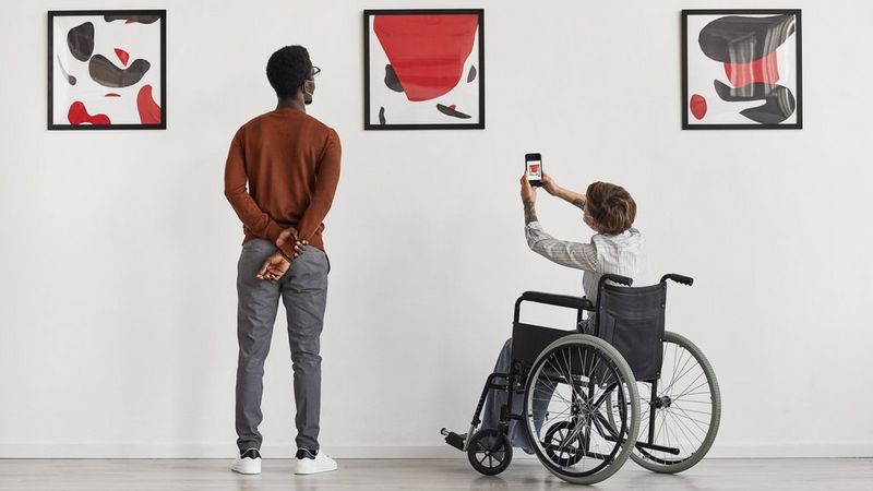 Aan de linkerkant kijkt een man met zijn handen achter zijn rug naar een muur waar drie rood-zwarte abstracte afbeeldingen hangen. Rechts van hem kijkt een man in een rolstoel via zijn telefoon naar een van de schilderijen.