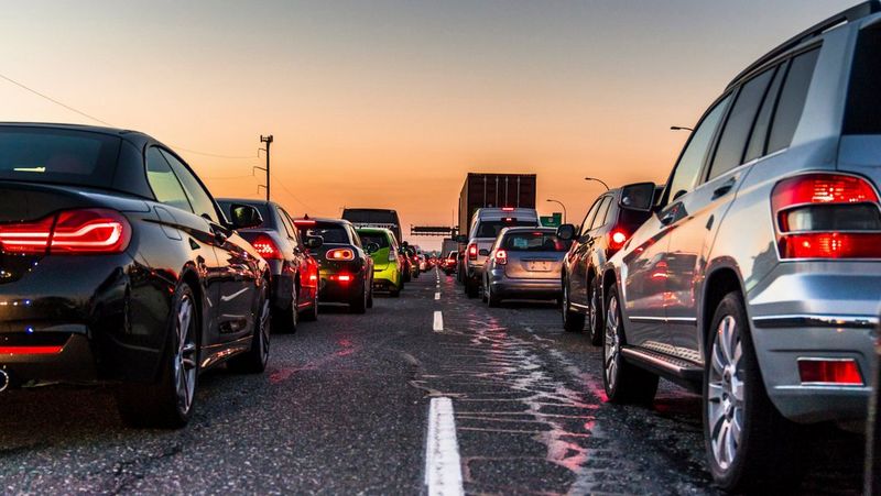 A photo taken from the centre of a traffic jam, standing on the central white live, viewing cars on left and right from behind. It is sunset and both cars and lorries are at a standstill, reflecting each other and their lights in their paintwork. In the distance there are power lines connected to telegraph poles. 