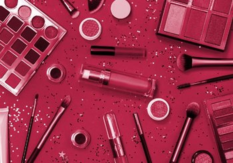 A selection of cosmetics and make-up brushes lying on a crimson/magenta table. There are also sparkles and glitter on the table.