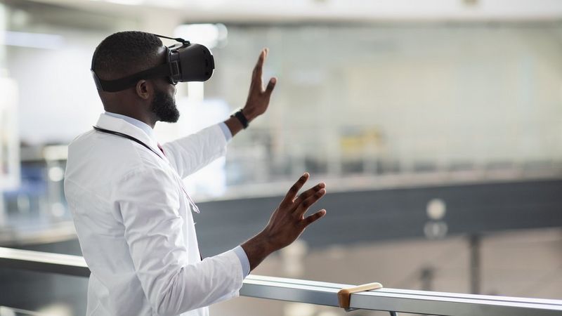 A doctor wearing a white lab coat and a stethoscope interacts with an invisible interface while wearing a virtual reality (VR) headset. He is standing indoors near a railing, with his hands raised in front of him as if manipulating a virtual environment.