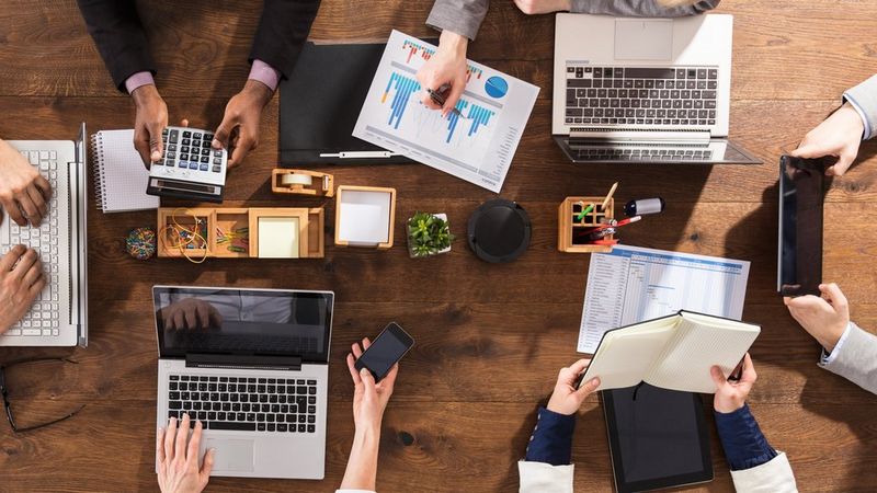 Vijf handen, laptops, telefoons en diverse kantoorartikelen en papieren op een tafel, van bovenaf bekeken.