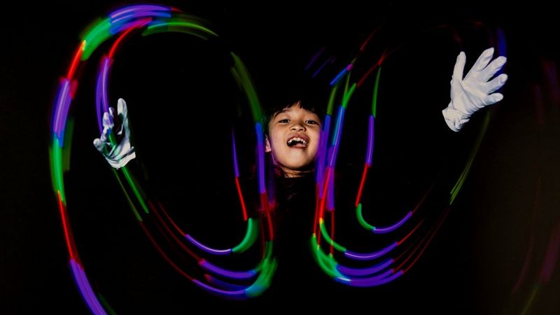 Vor einem schwarzem Hintergrund sieht man das Gesicht eines lachenden japanischen Kindes, seine weiß behandschuhten Hände und Lichtspuren in Grün, Lila und Rot.